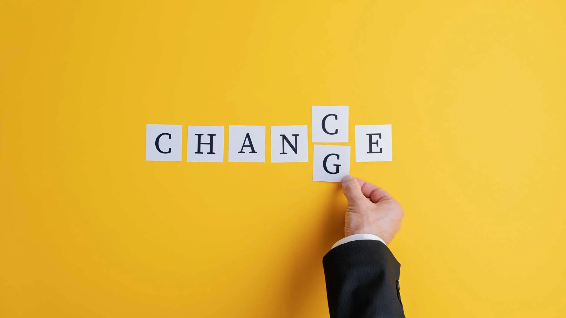 A hand rearranges letter cards spelling “CHANGE” into “CHANCE” against a bright yellow background.