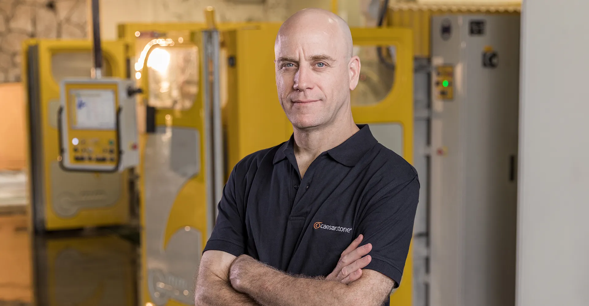 Fabricator at Caesarstone’s Master of Stone Training Center standing confidently in front of machinery, wearing a Caesarstone-branded shirt inside a professional workshop.